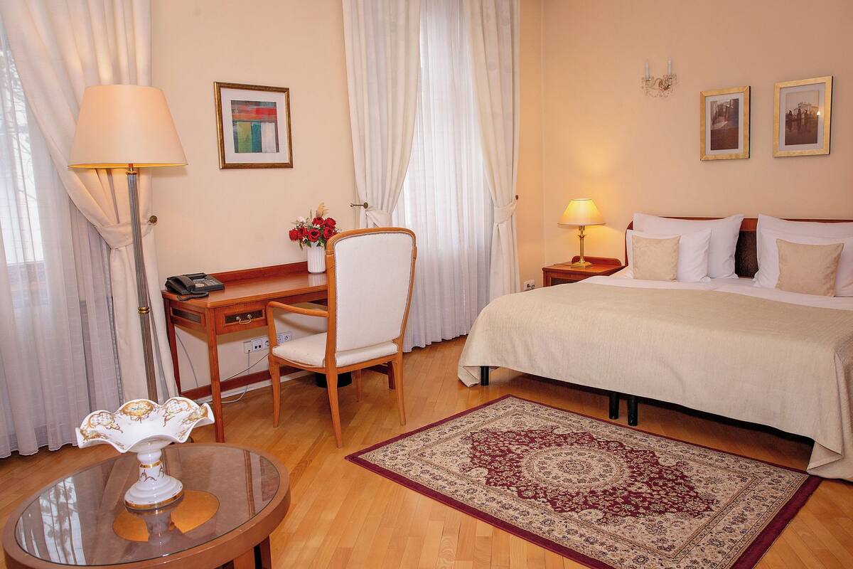 A hotel room with a bed, a desk with a chair, a lamp, a vase with flowers, and a rug.