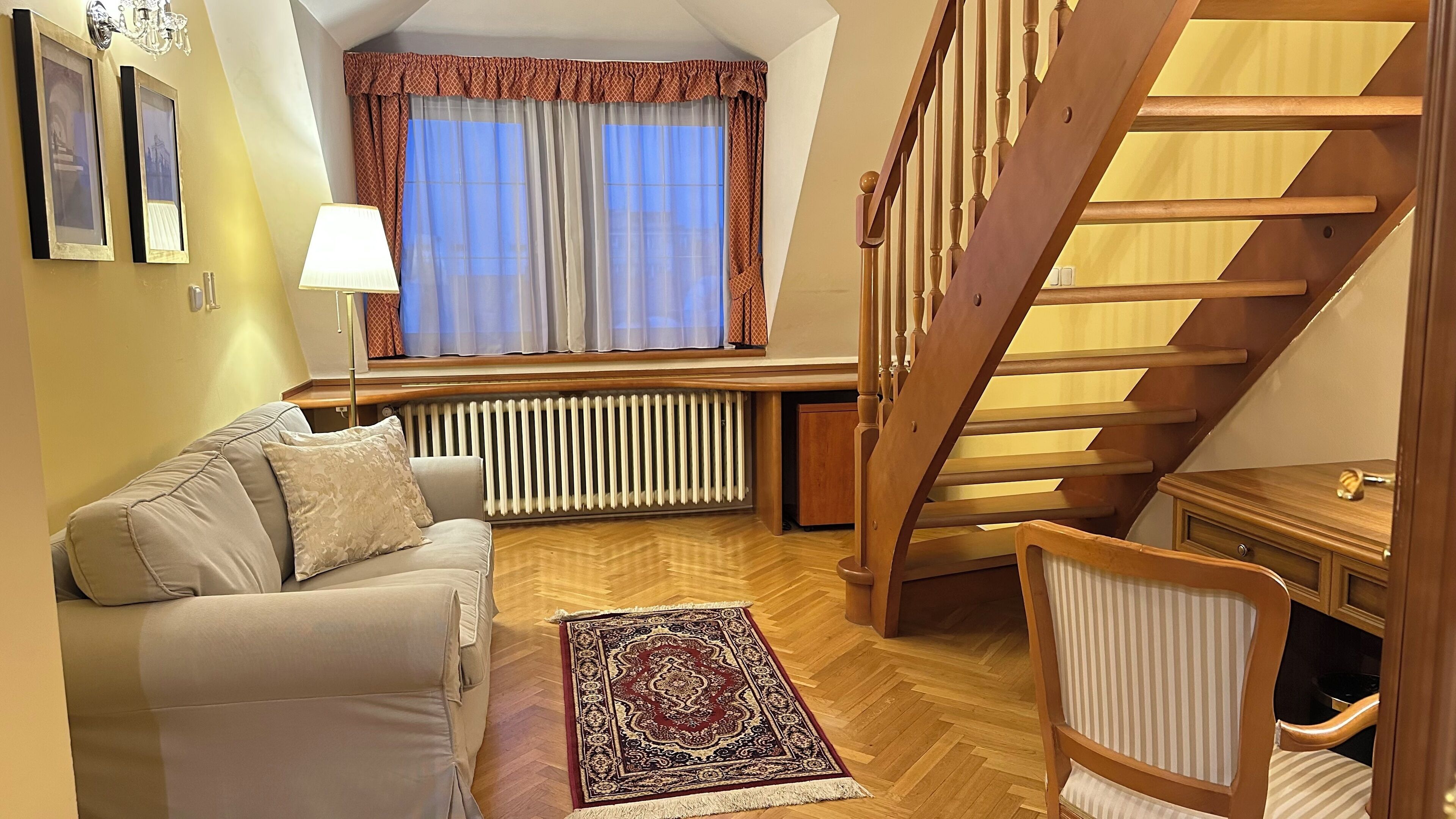 Una sala acogedora con un sofá, un tapete estampado, una silla de madera, un radiador y una escalera.