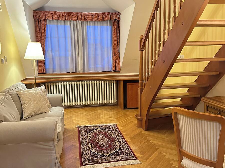 Una sala acogedora con un sofá, un tapete estampado, una silla de madera, un radiador y una escalera.