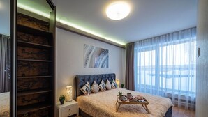 Een moderne slaapkamer met een groot bed, een nachtkastje met lamp, een dienblad met ontbijtartikelen en uitzicht buiten door dunne gordijnen.