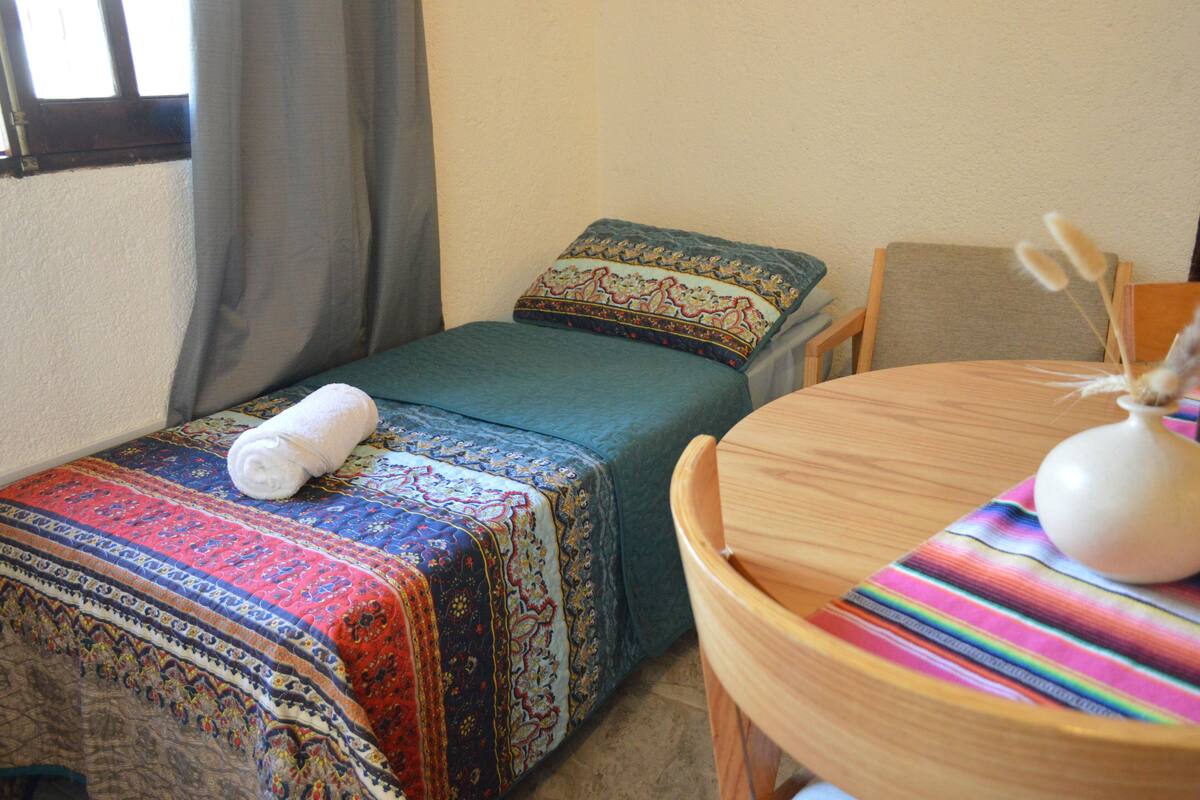 Una cama pequeña y bien hecha con un edredón colorido y una toalla enrollada. Hay una silla de madera y una mesita con un jarrón y un objeto decorativo.