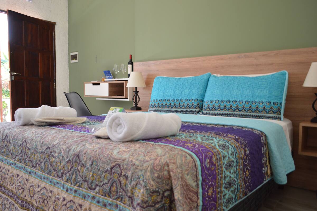 Una cama bien hecha con una colcha estampada, dos almohadas con un diseño geométrico, una mesita de noche con una lámpara y una mesita auxiliar de madera.