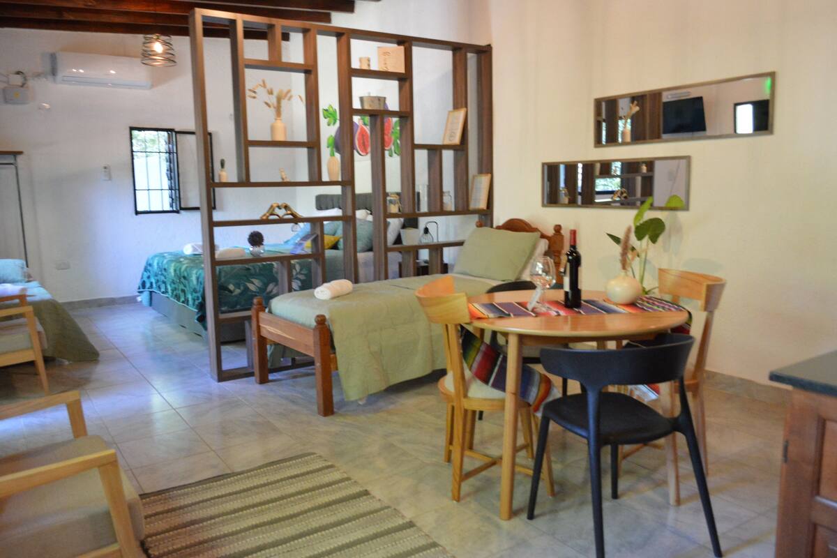Un salón acogedor con una cama, una mesa de comedor con sillas, un separador de madera con obras de arte y una pequeña cocina.
