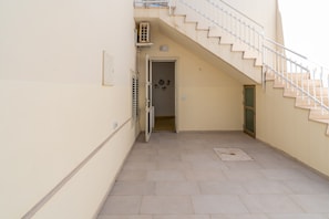 Un corredor estrecho con una escalera que sube a un piso superior, una puerta con celosía metálica y un suelo de baldosas.