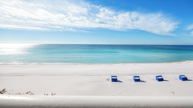 Ein Strand mit weißem Sand und einem strahlend blauen Himmel. Drei blaue Liegen stehen in einer Reihe im Sand.