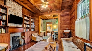 A cozy log cabin interior with a fireplace, bookshelves, a television, and a dining area.