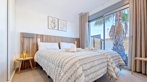 A modern hotel room with a large bed, a bedside table with a lamp, a view of palm trees through large windows, and a wooden headboard. - Villa with private pool, roof terrace and BBQ (Los Alcazares)