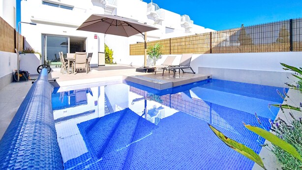 A modern pool area with a clear blue pool, a covered seating area with chairs and an umbrella, and a white building with balconies in the background. - Villa with private pool, roof terrace and BBQ (Los Alcazares)