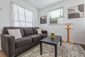 Una sala de estar con un sofá gris oscuro, una mesita con una planta, un perchero de madera y una ventana con cortinas.