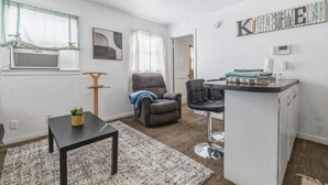 Ett kompakt bostadsutrymme med ett litet pentry, en grå fåtölj, ett litet bord och en krukväxt.