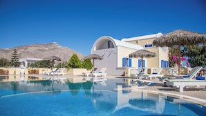 Una piscina azul cristalina con sillas de descanso y sombrillas, rodeada de edificios blancos con detalles azules y una montaña al fondo.