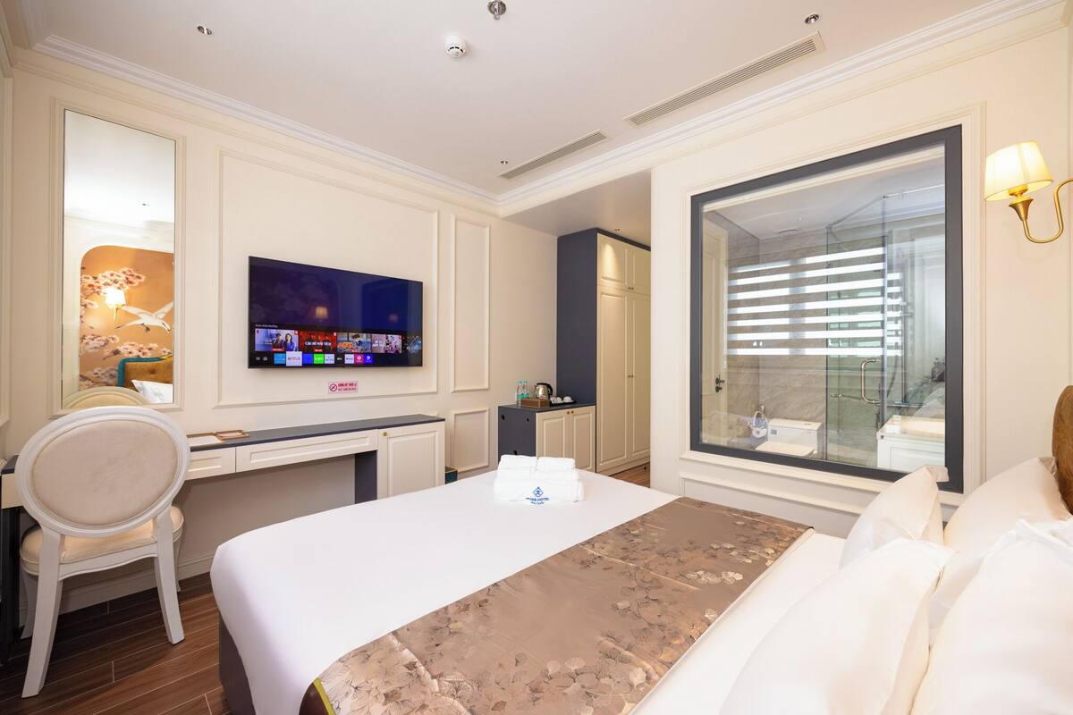 A hotel room with a bed, a desk, a chair, a television, and a bathroom with a glass door.