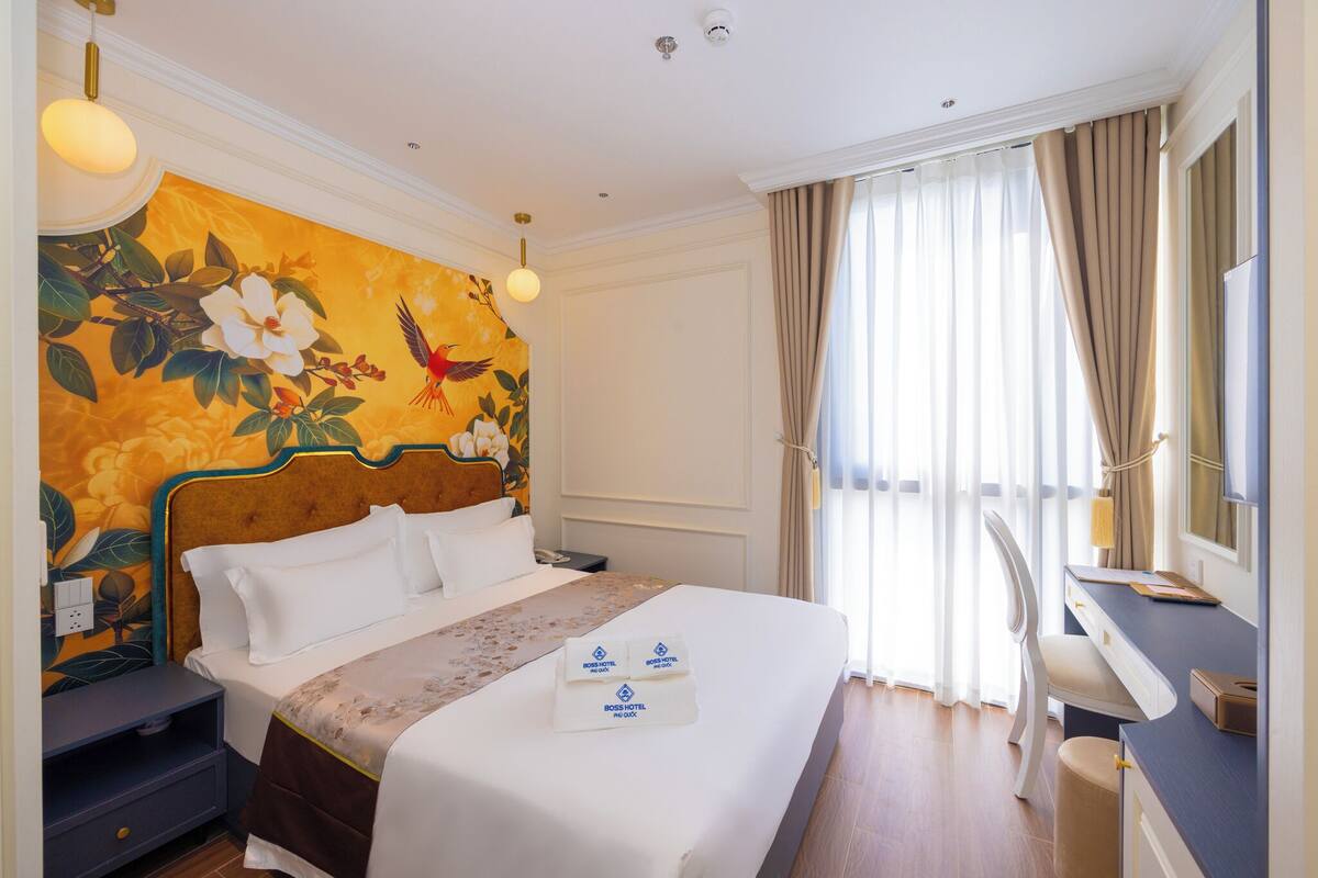 A hotel room with a large bed, a desk, and a chair. There is a painting on the wall, a window with curtains, and a ceiling light.
