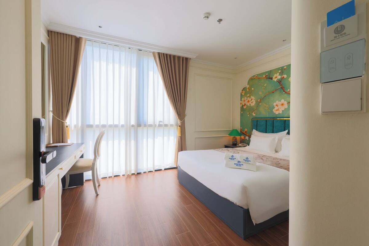 A hotel room with a bed, a desk, and a chair. There is a window with curtains, a wall-mounted air conditioner, and a floral wall decoration.
