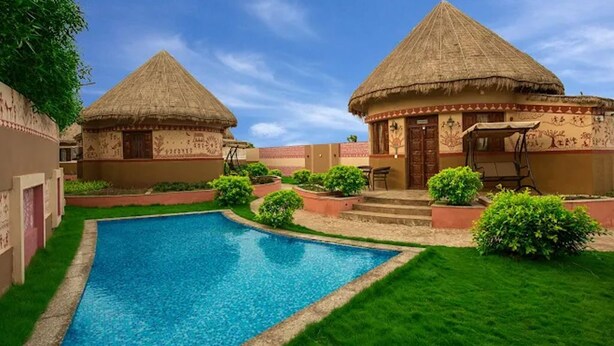 A pool area with two thatched-roof huts, surrounded by well-maintained lawns and small bushes. - The Sky Imperial Chotila (Chotila)