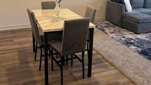 Een eetgedeelte met een houten tafel en stoelen, een vaas met gedroogde planten, een grijze bank en een vloerkleed met patroon.