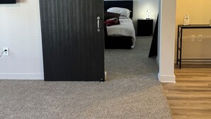 Een slaapkamer met een donker houten deur, een bed met wit beddengoed, een nachtkastje en een kunstwerk aan de muur.