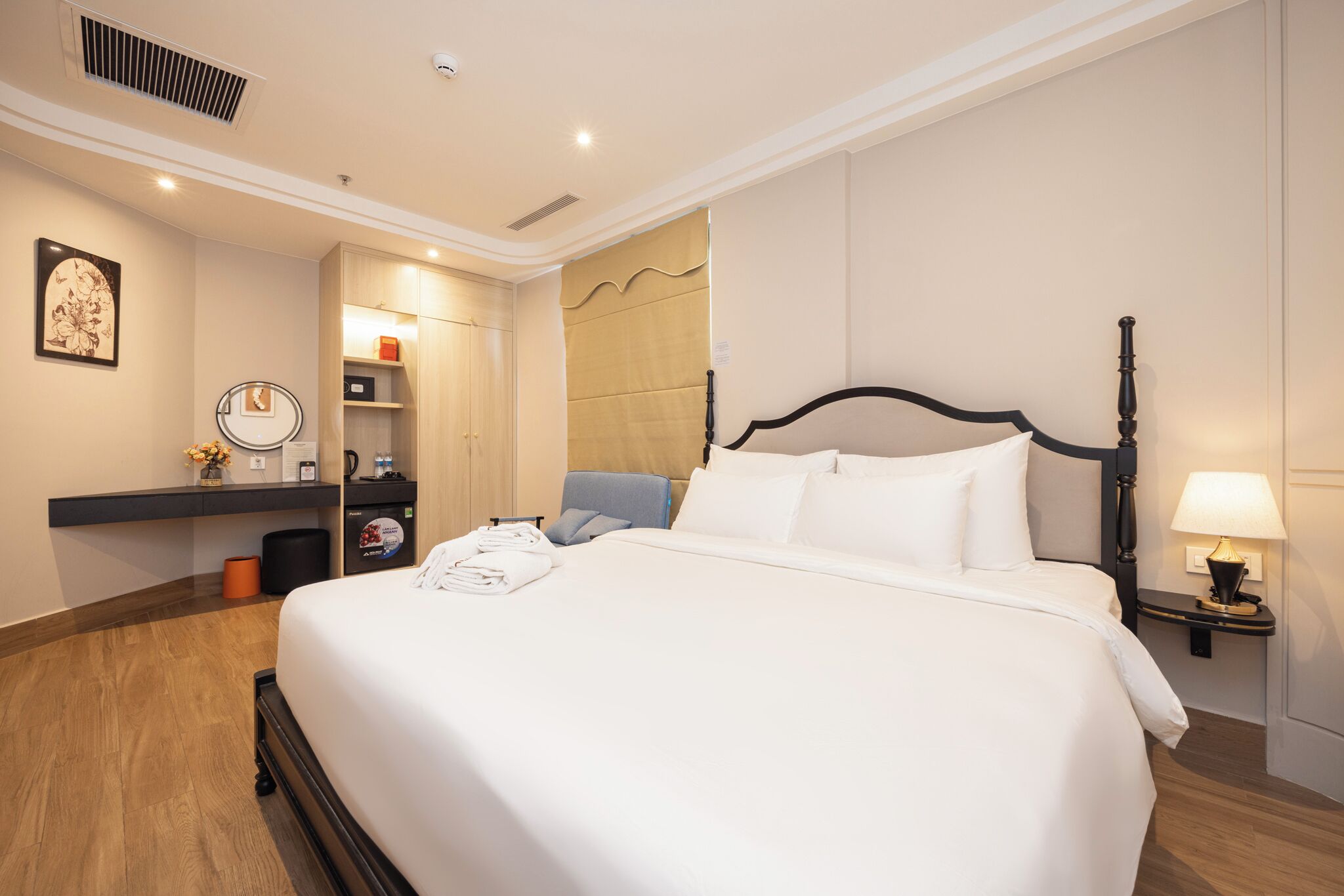 Una habitación de hotel con una cama grande, una mesita de noche con lámpara, una silla, un escritorio con espejo y un estante con artículos de aseo.