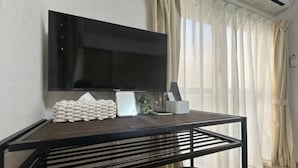 Una habitación con una televisión de pantalla plana montada en la pared, una mesa de madera con una silla y una ventana con cortinas translúcidas.