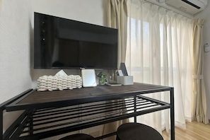 Una habitación con una televisión de pantalla plana montada en la pared, una mesa de madera con una silla y una ventana con cortinas translúcidas.