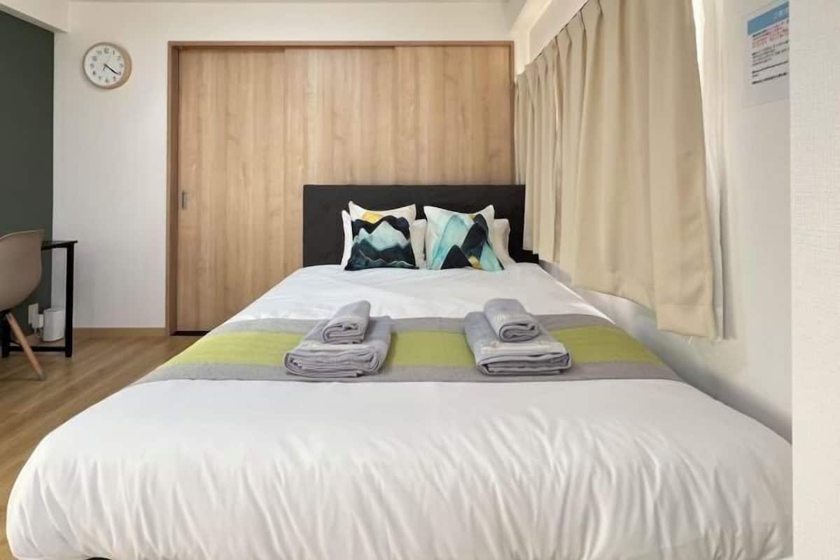 Una cama bien tendida con dos toallas dobladas, un reloj en la pared y un armario de madera.