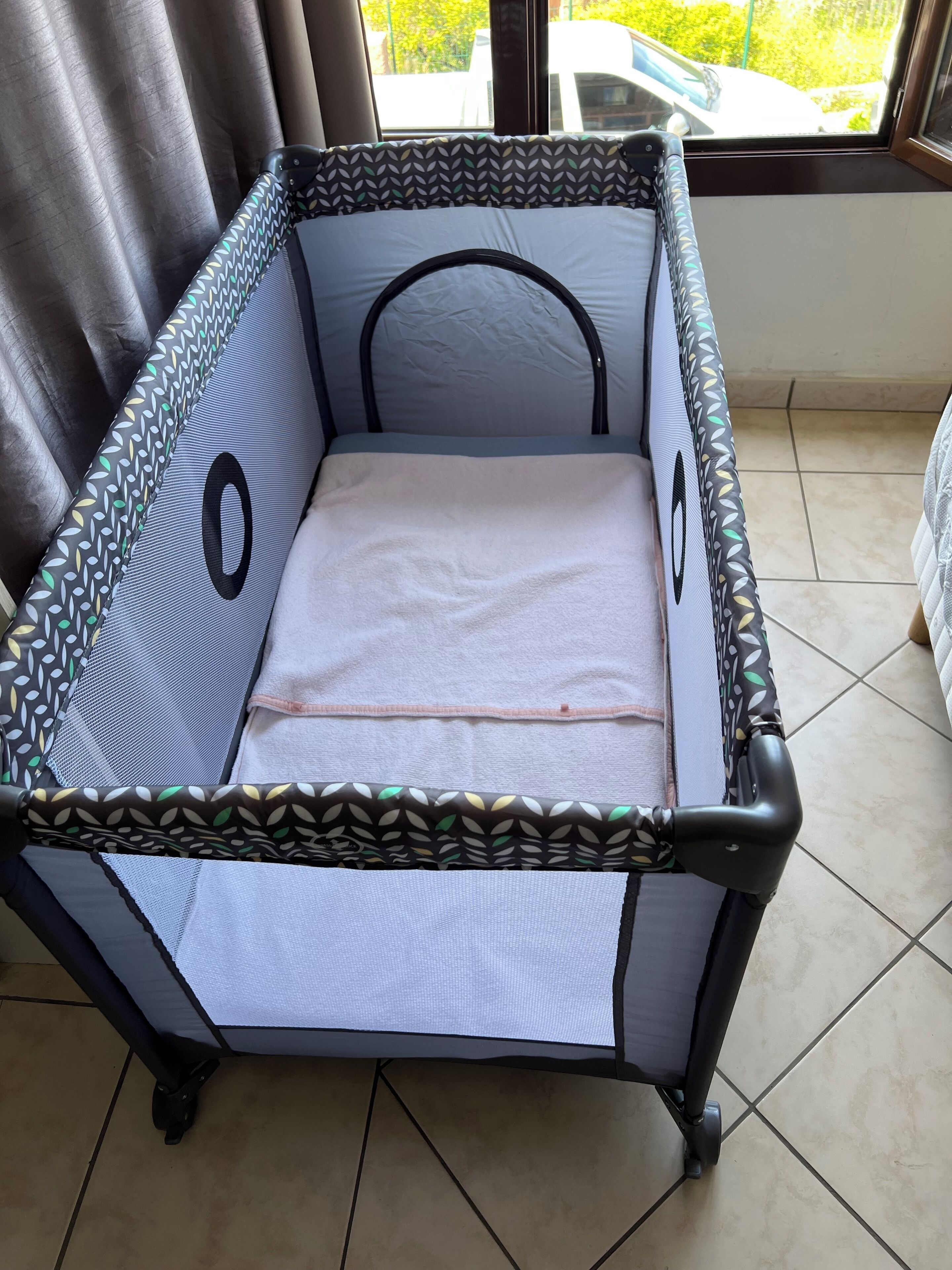 Un berceau pour bébé avec un bord décoré, un matelas blanc et une vue sur une voiture garée, visible à travers une fenêtre.
