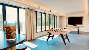 A modern living room with a foosball table, a television, and large windows offering a view of the outdoors. - Ferme Valentour 24 beds, ideal for families and corporate seminars. (Gesves)