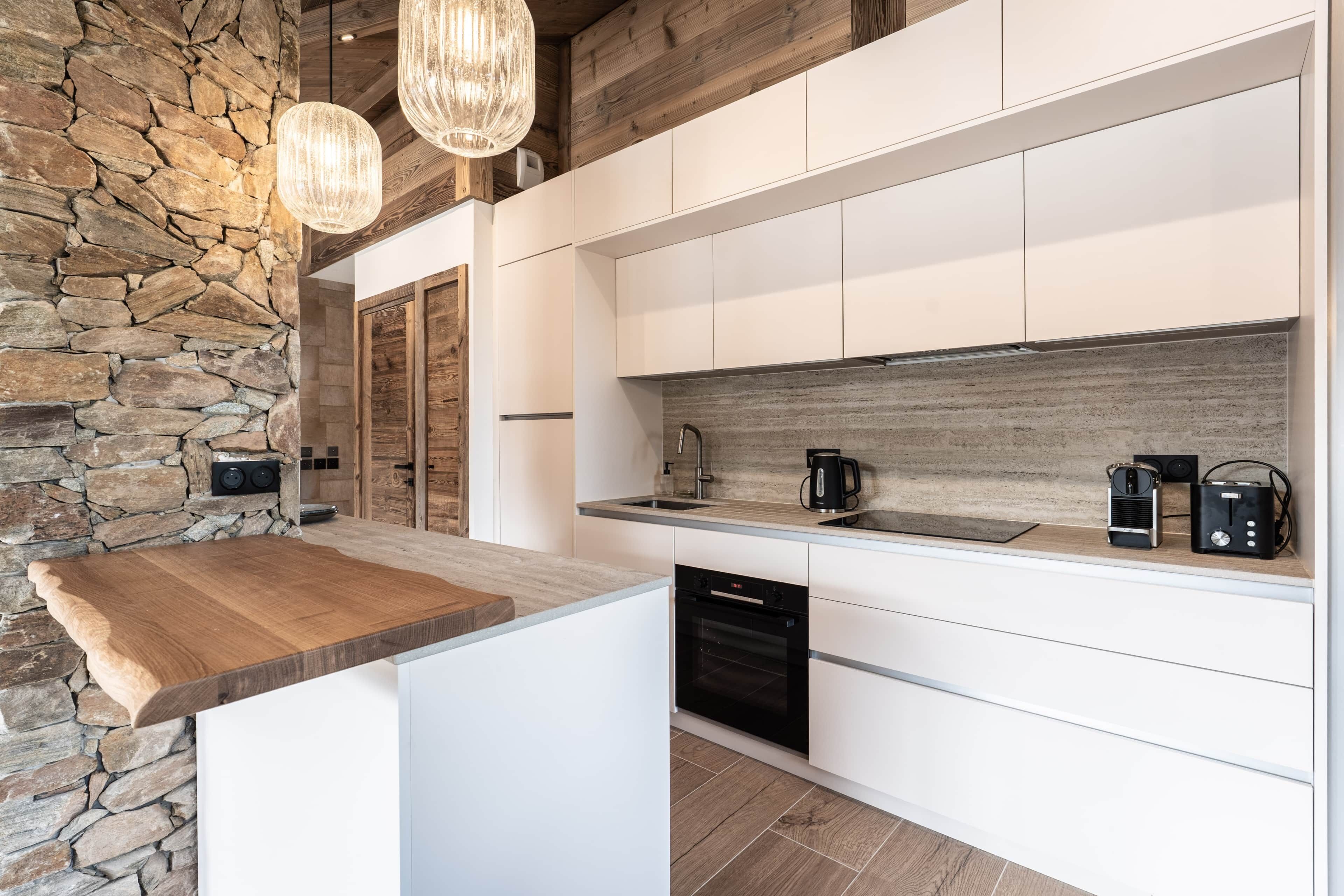 A modern kitchen with a stone accent wall, wooden ceiling beams, and white cabinetry.