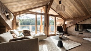 A spacious loft with a high ceiling, large windows offering mountain views, a cozy seating area with a sofa and armchairs, and a flat-screen TV mounted on the wall. - Penthouse in Courchevel Moriond, Belvédère area (Courchevel)