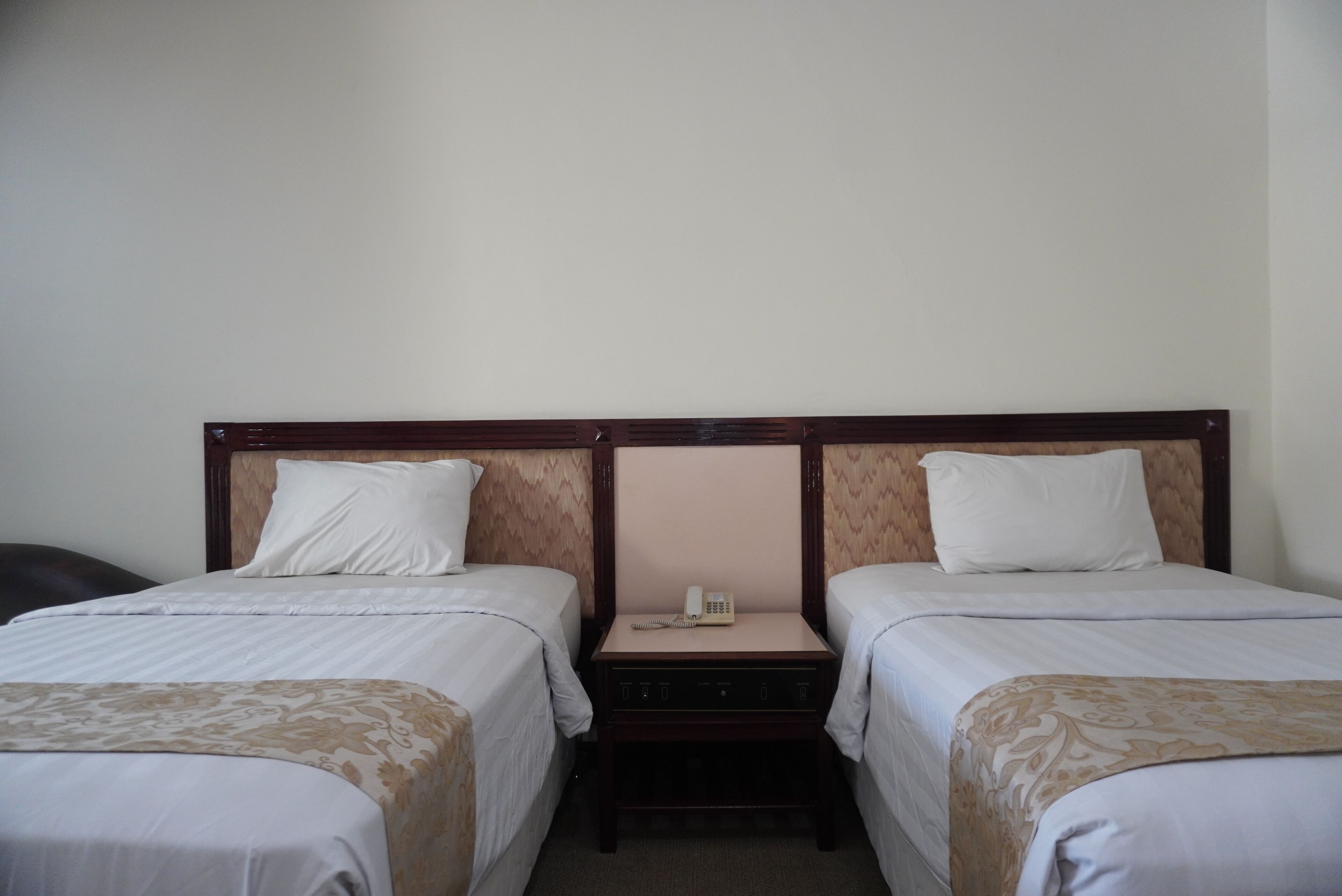 Une chambre d’hôtel avec deux lits simples, une tête de lit en bois, une table de chevet avec un téléphone et un petit tiroir.