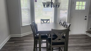 Een eetruimte met een ronde tafel en zes stoelen, grote ramen met zonwering, een kroonluchter en een houten vloer.