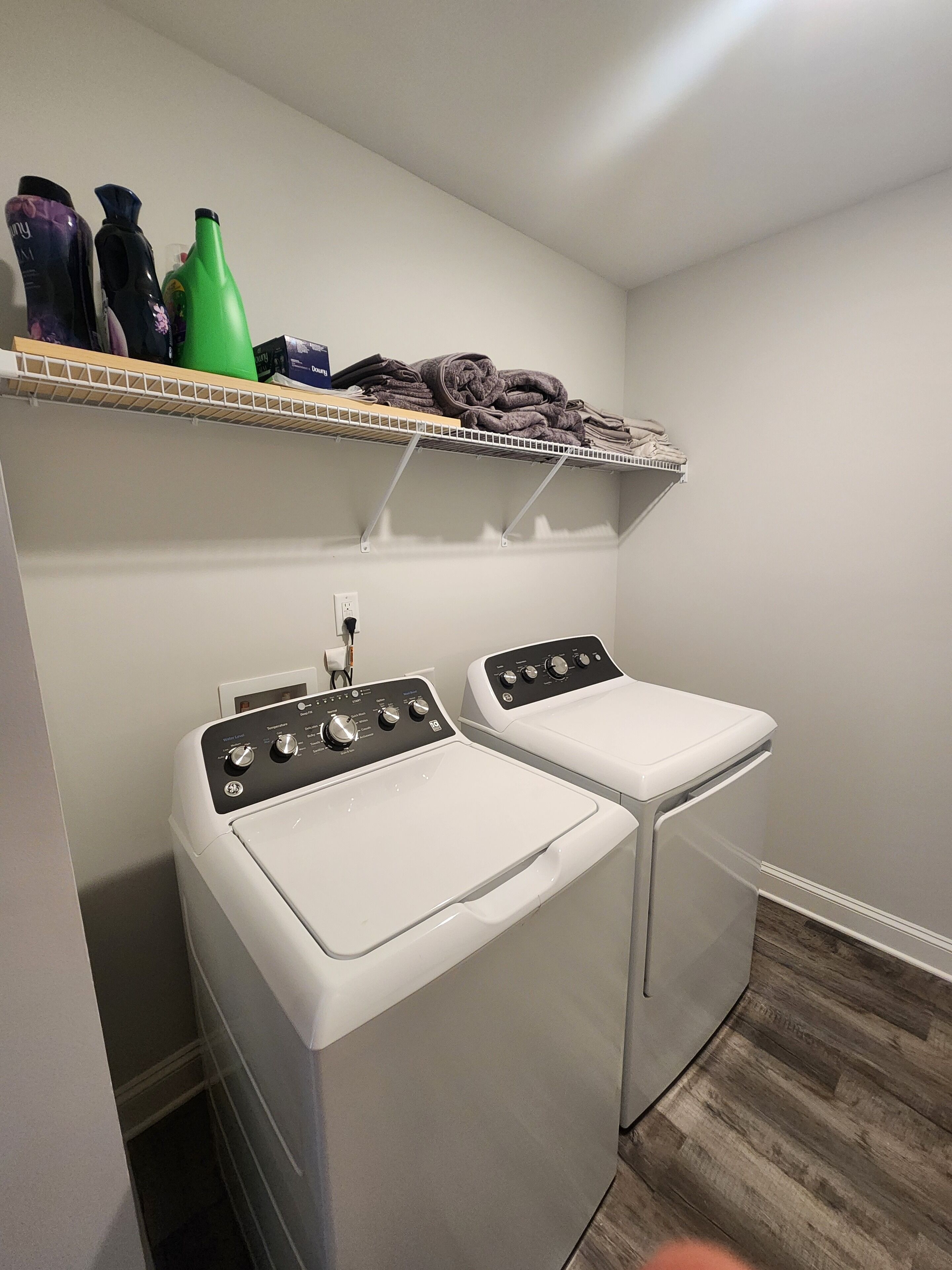 A laundry area with two white front-loading washing machines and a shelf above holding folded towels and cleaning supplies.