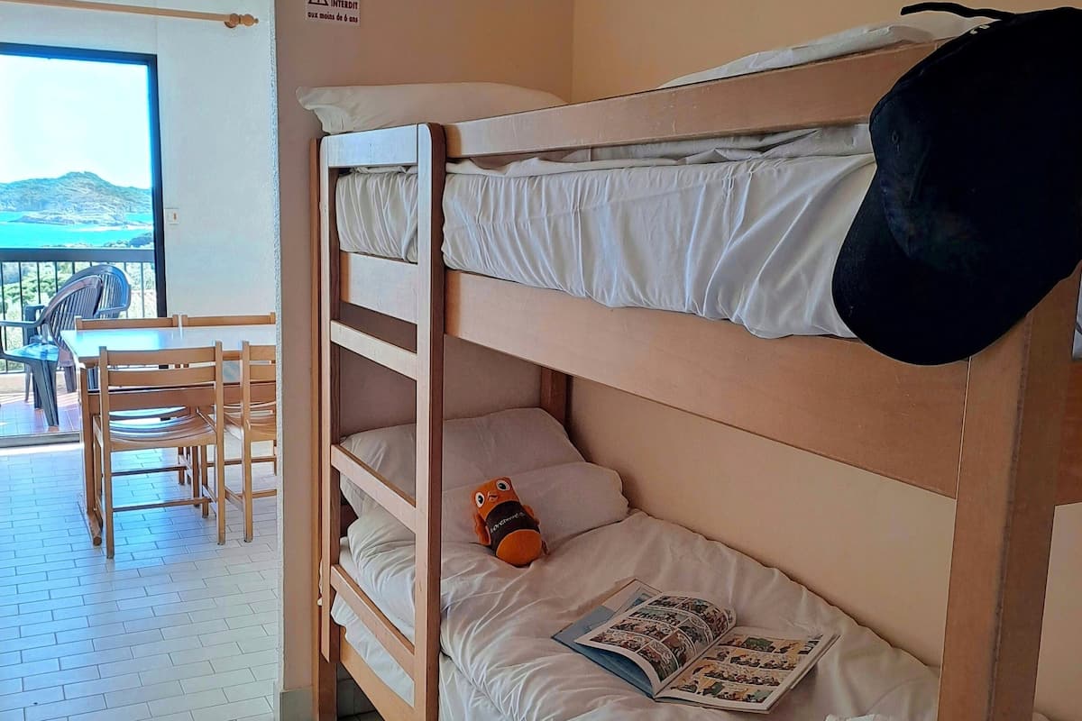 A bunk bed with a pillow and a book, a table with chairs, and a view of the sea.