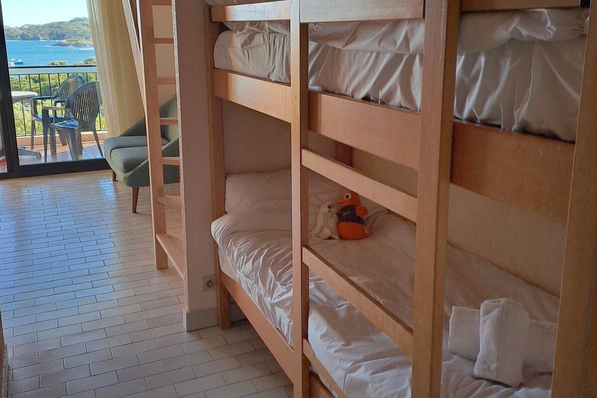 A bunk bed room with a view of the sea, a balcony with chairs, and a wooden floor.