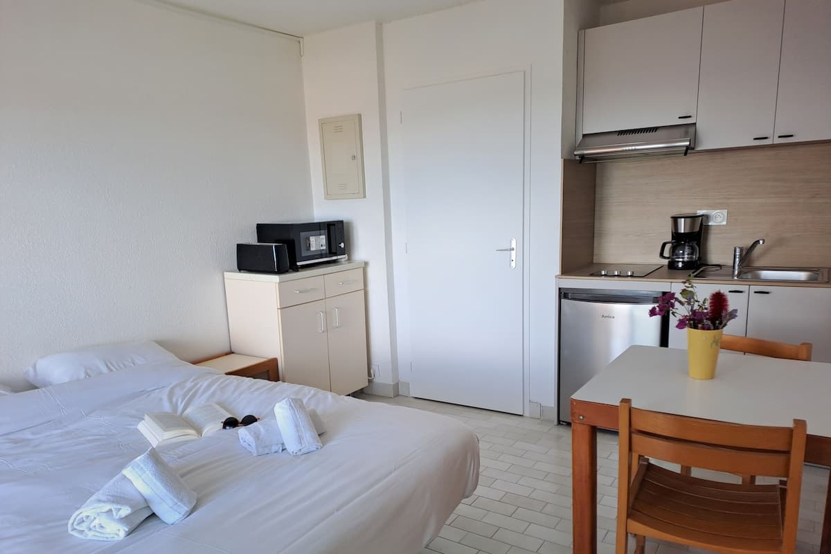 A hotel room with a bed, a desk with a chair, a kitchenette, and a bathroom door.