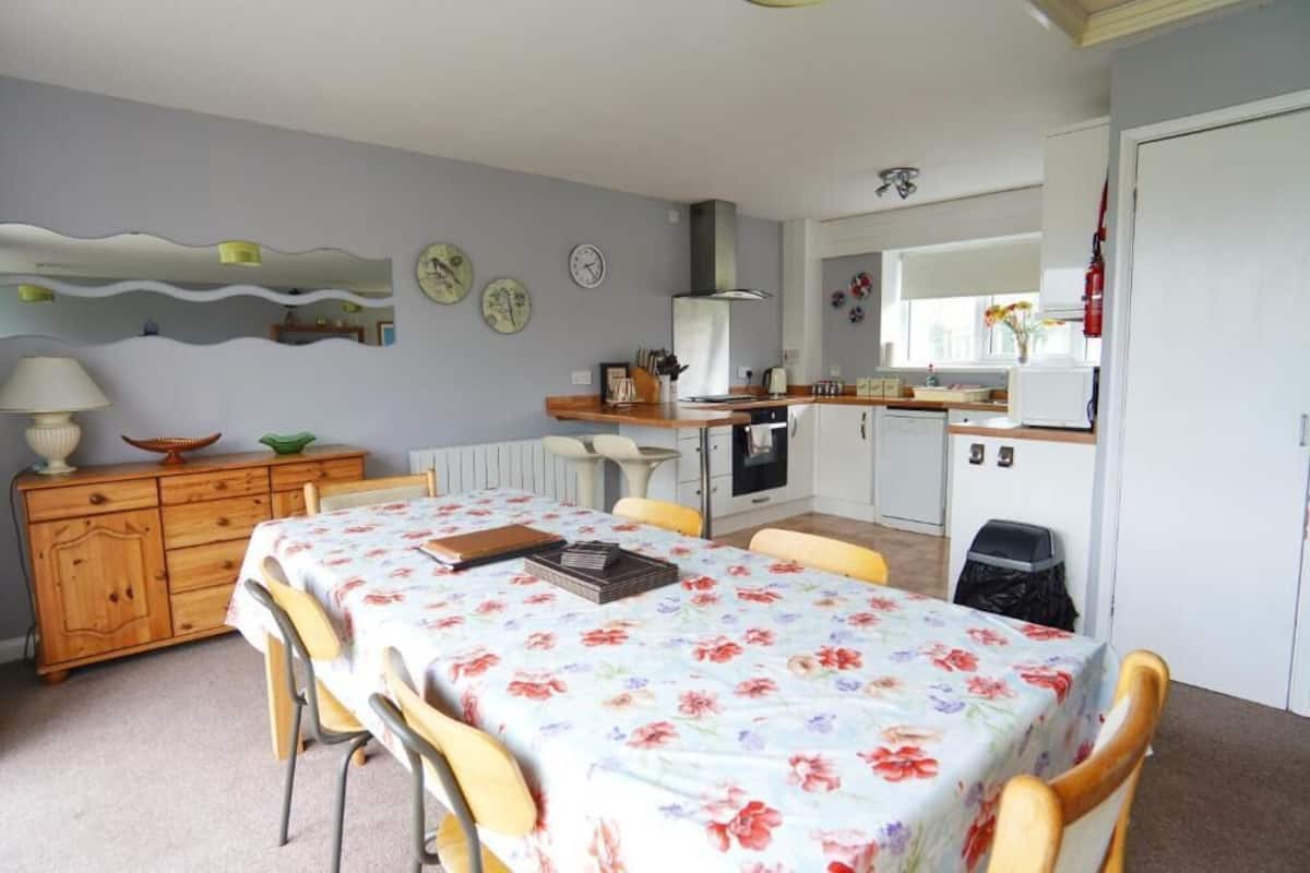 Ein Essbereich mit einer blumigen Tischdecke, Holzsesseln, einem Holzschrank und einer Küche im Hintergrund.