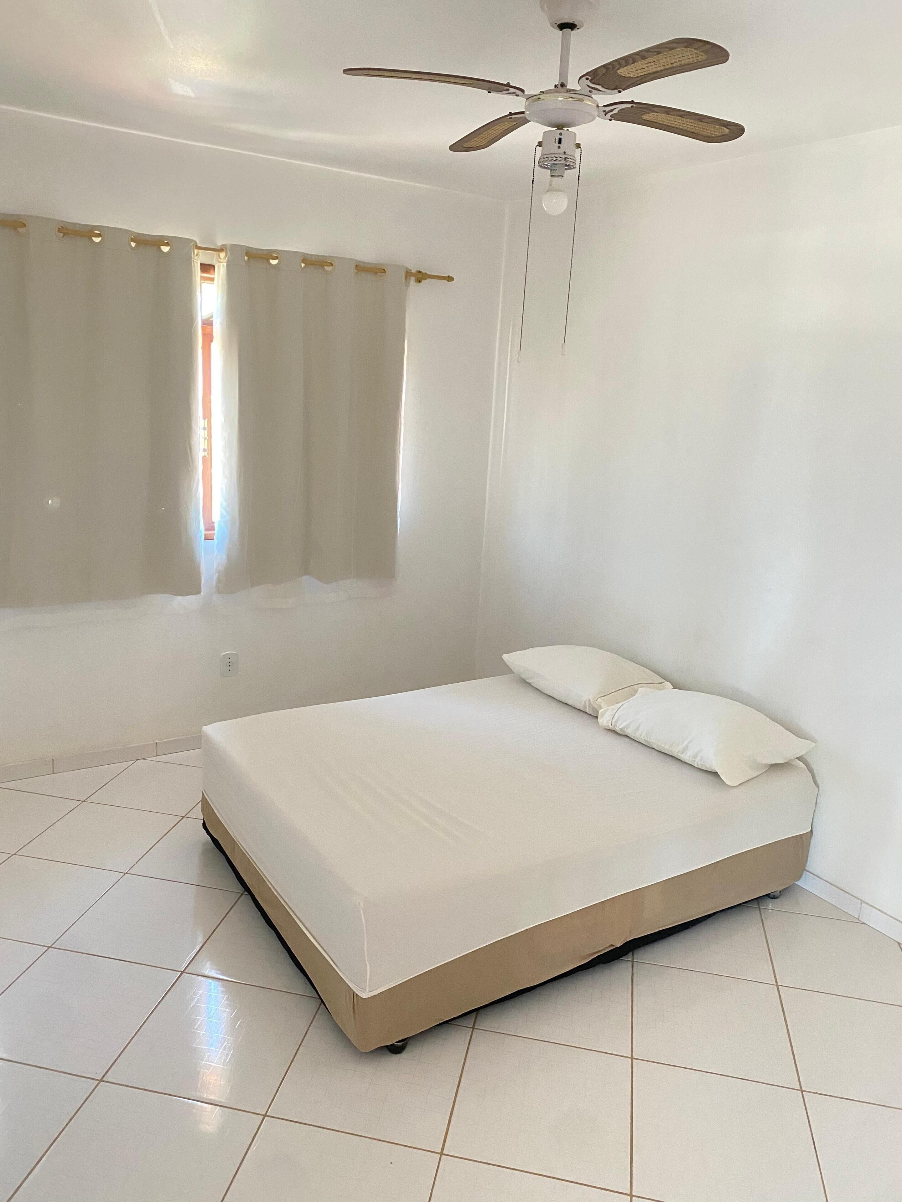 Een slaapkamer met een bed, witte gordijnen, een plafondventilator en een raam.