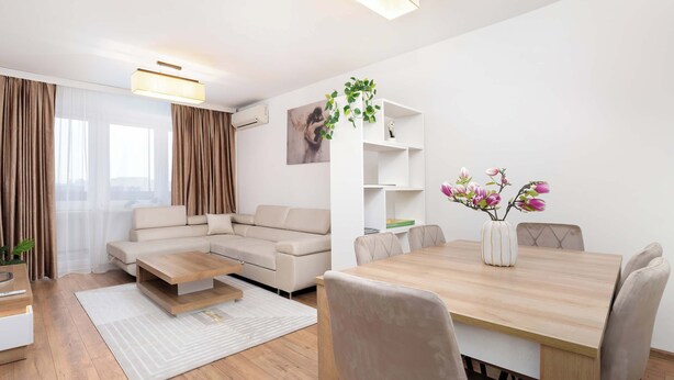 A modern living room with a sofa, coffee table, and dining table with chairs. There is a white wall with a painting and a plant on a shelf. - Family Apartment in the Center | 3 Bedrooms and Air Conditioning (Rzeszów)
