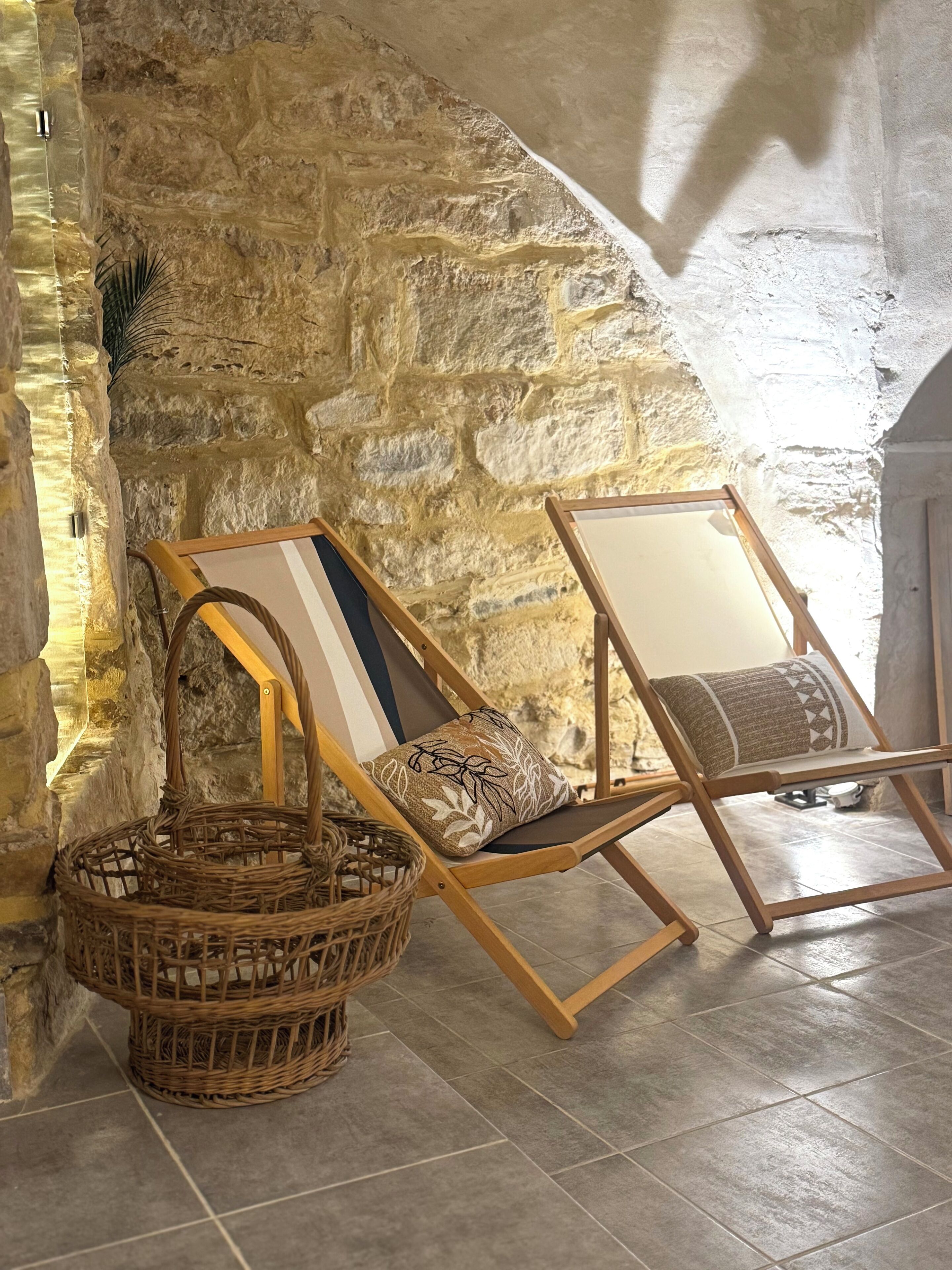Two wicker chairs with cushions against a stone wall, a woven basket, and a tiled floor.