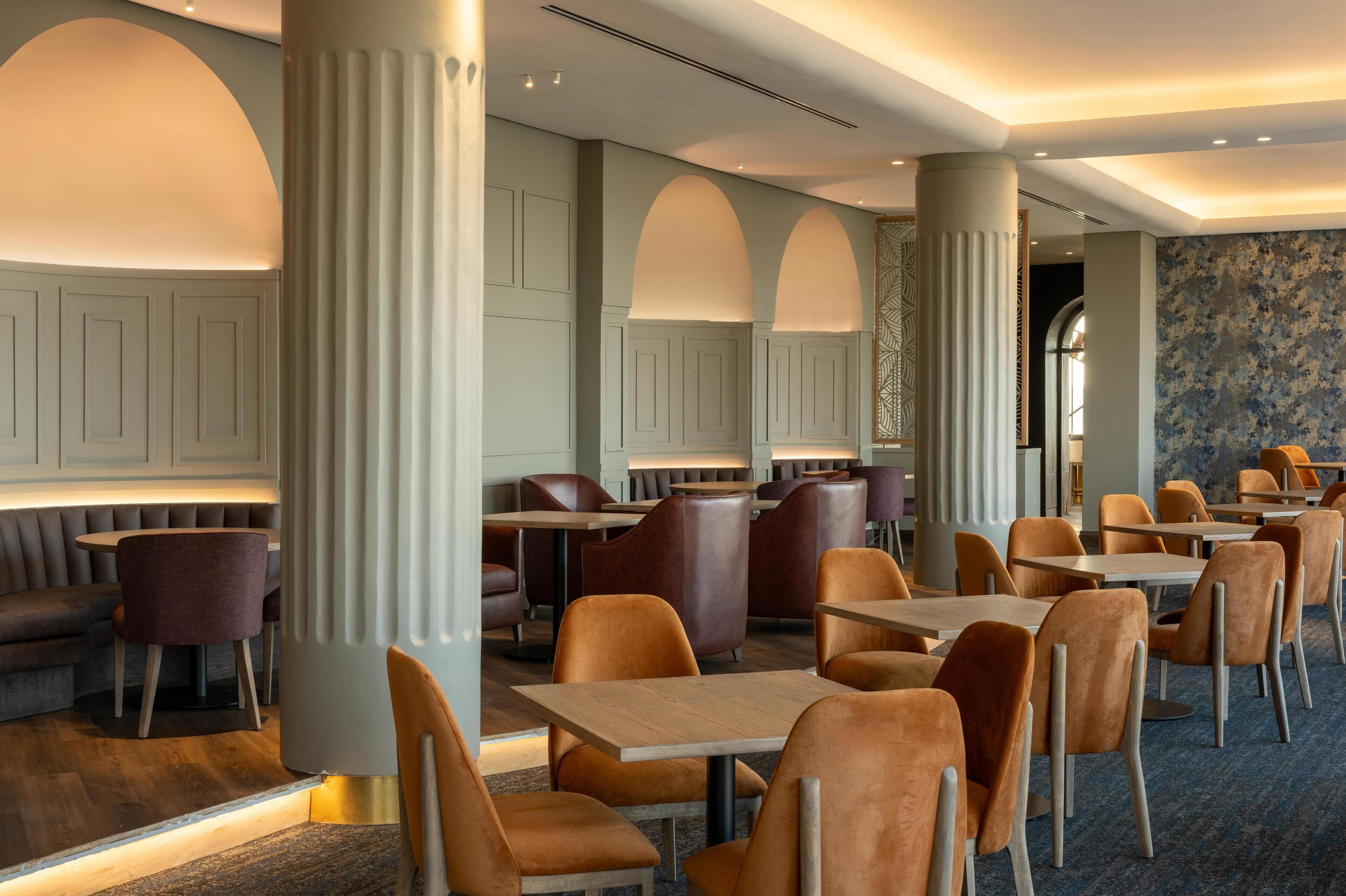 A modern dining area with high tables and cushioned chairs, featuring a mix of wood and leather finishes.