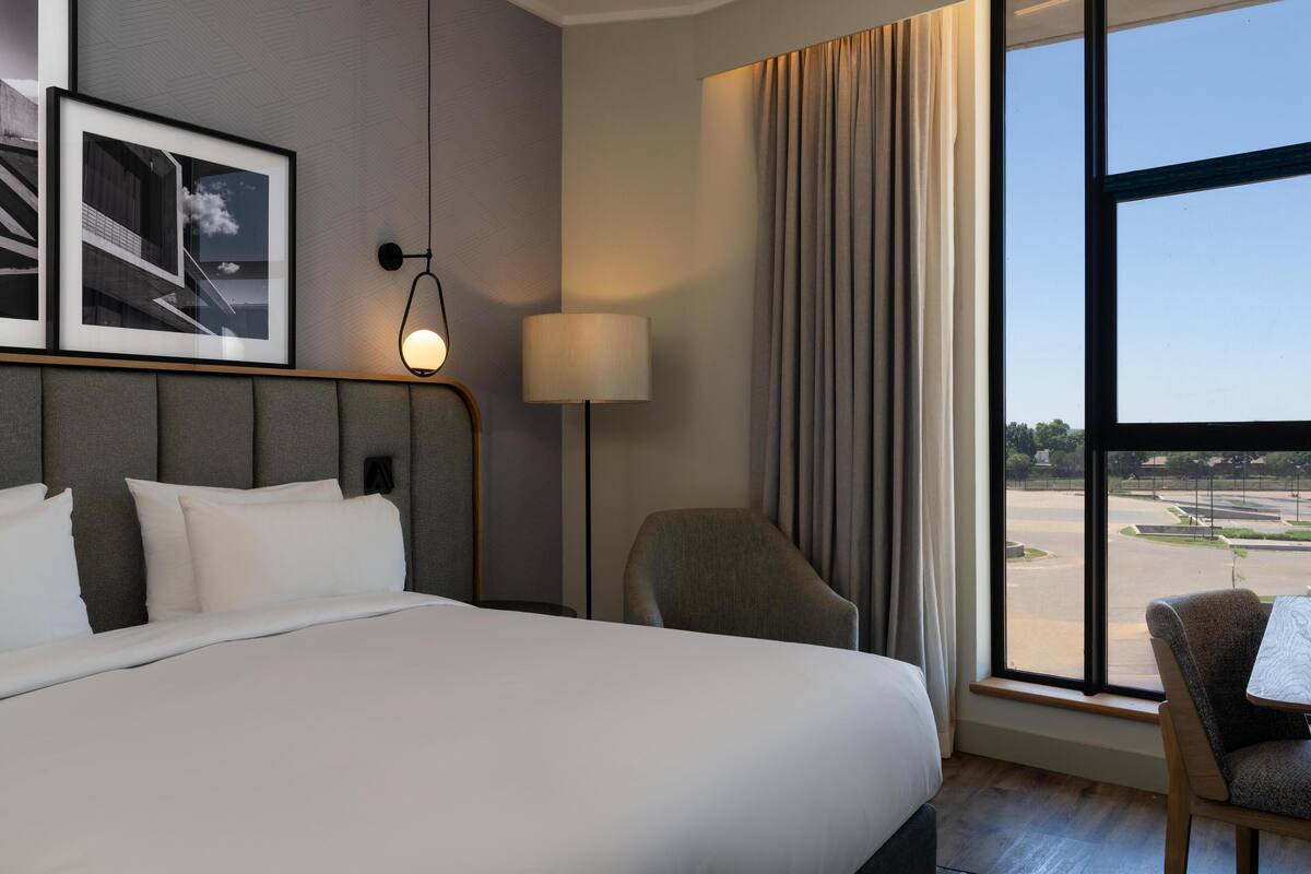 Habitación de hotel con una cama grande, una silla, una lámpara, un cuadro enmarcado y vistas al exterior.