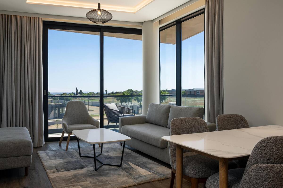 Una sala de estar moderna con un sofá, sillas, una mesa y ventanas amplias que dan a un paisaje exterior.