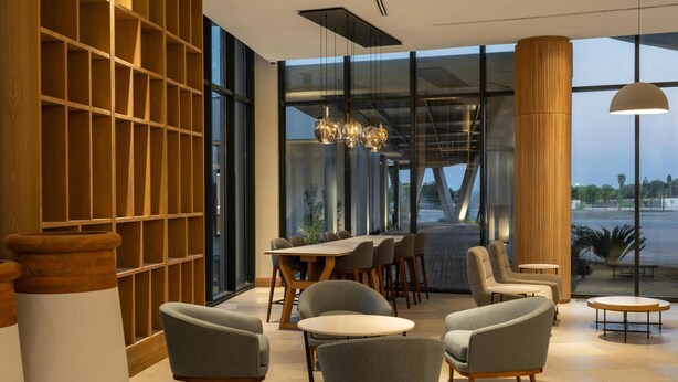 A modern indoor space with a large window, a wooden bookshelf, and a seating area with round tables and chairs. - Radisson Hotel And Convention Center Steve Tshwete Middelburg (Middelburg)