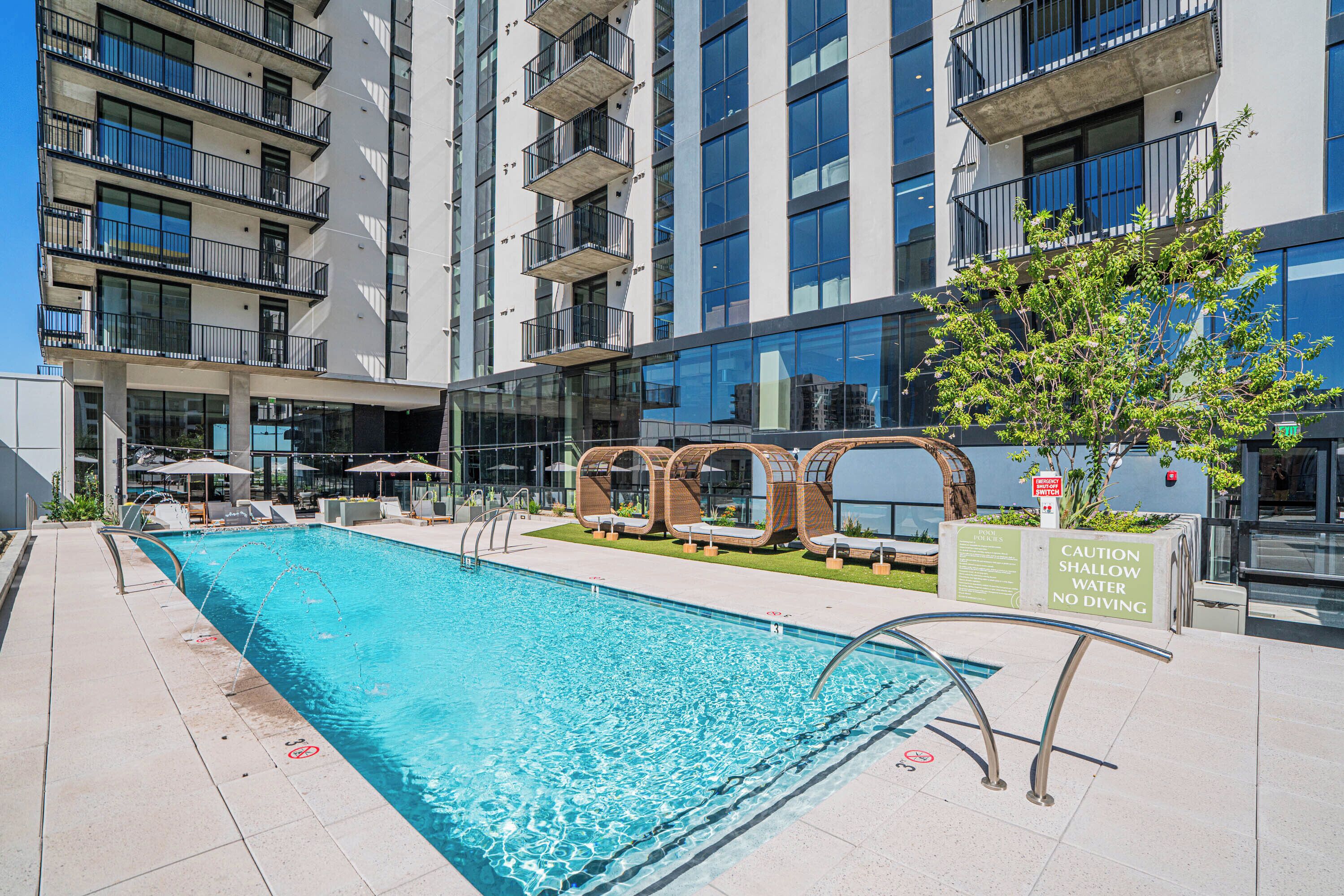 A swimming pool with lounge chairs and a caution sign, surrounded by a modern building with balconies.