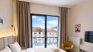 Quarto de hotel com uma cama grande, uma poltrona amarela, uma televisão e uma varanda com vista para edifícios.