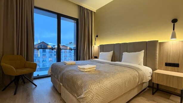 Quarto de hotel com uma cama grande, uma cadeira, uma mesinha de cabeceira e vista para prédios através de uma janela ampla.