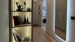 Um corredor com piso de madeira, uma estante com diversos objetos, uma máquina de lavar e um guarda-roupa espelhado.