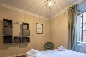 A hotel room with a bed, a chair, a wall-mounted shelf unit, a framed picture, and a window with curtains. - BuddyRent - Ghetto - Campo de' Fiori - 6 ospiti (Roma)