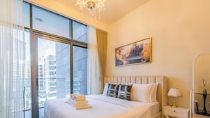 A modern hotel room with a large bed, a view of the city through floor-to-ceiling windows, a painting on the wall, and a bedside table with a lamp. - Tranquil Studio Getaway in Meydan City (Dubai)