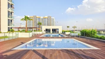 A rooftop pool area with a wooden deck, a swimming pool, and a building with multiple balconies in the background.